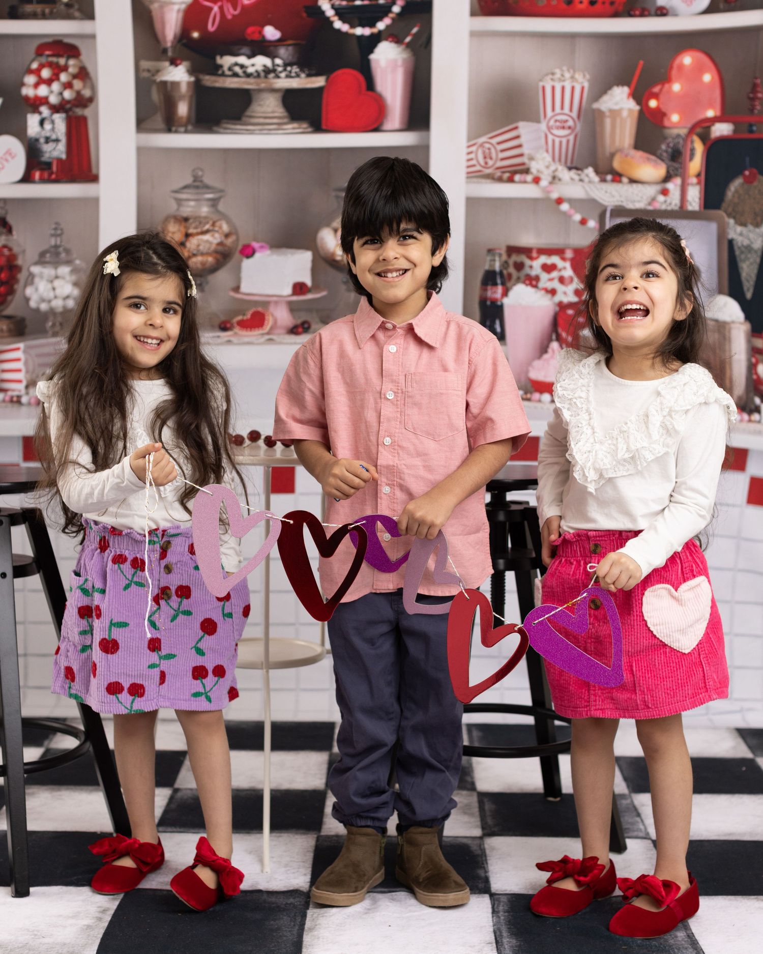 Children posing in Valentine's Day themed outfits with pink and red colors against a decorated backdrop.