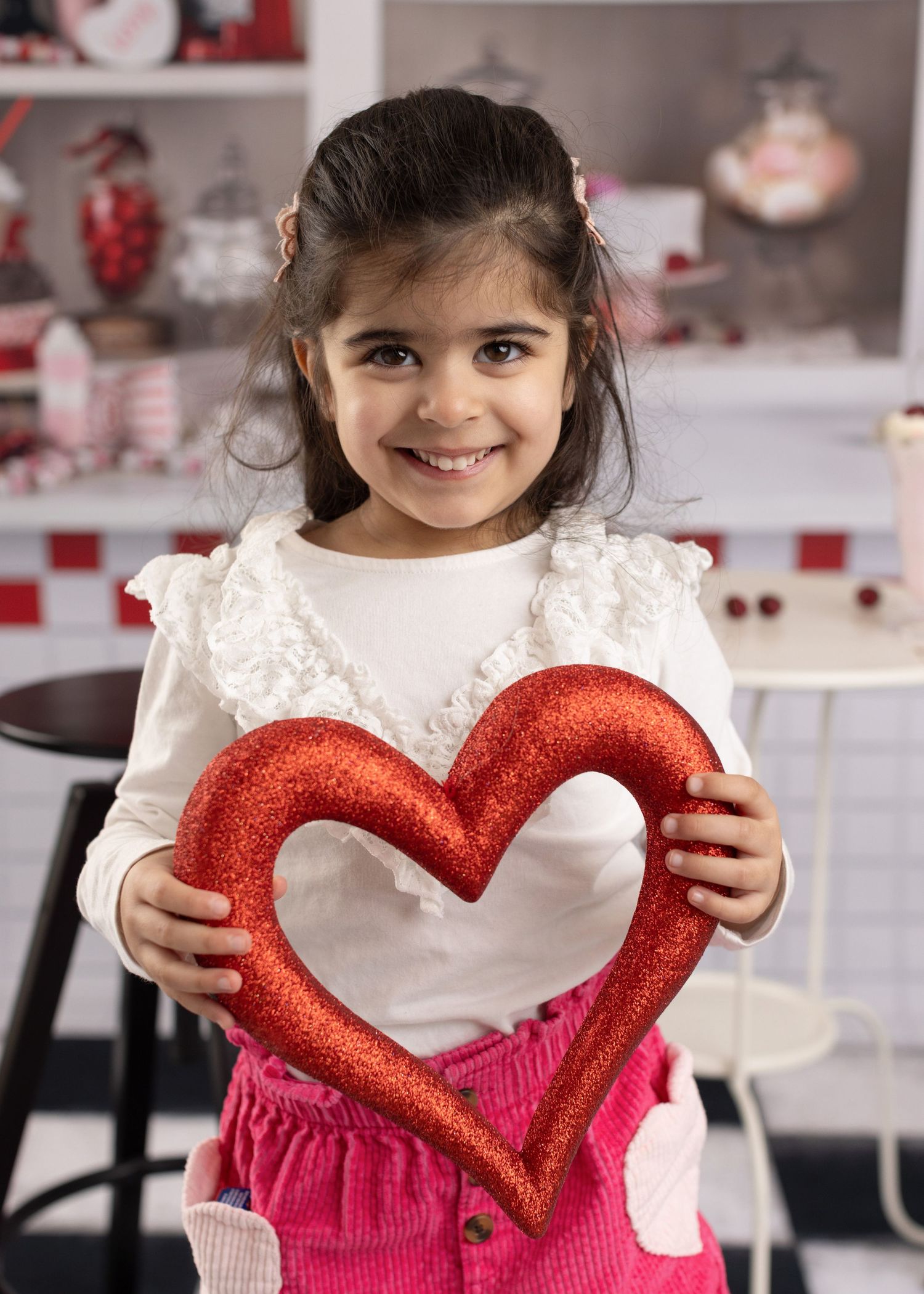 A child holds a sparkly red heart decoration while wearing a white long-sleeved dress in a festive holiday setting.