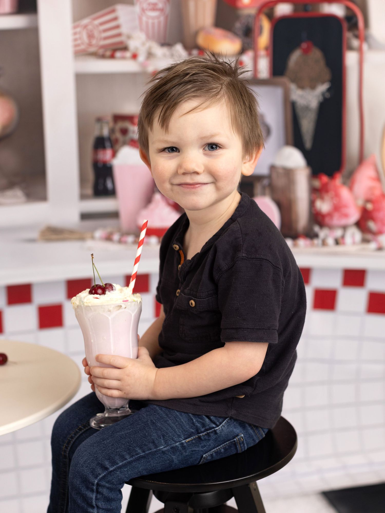 A child in a black shirt enjoys a pink milkshake in a retro-styled diner setting.