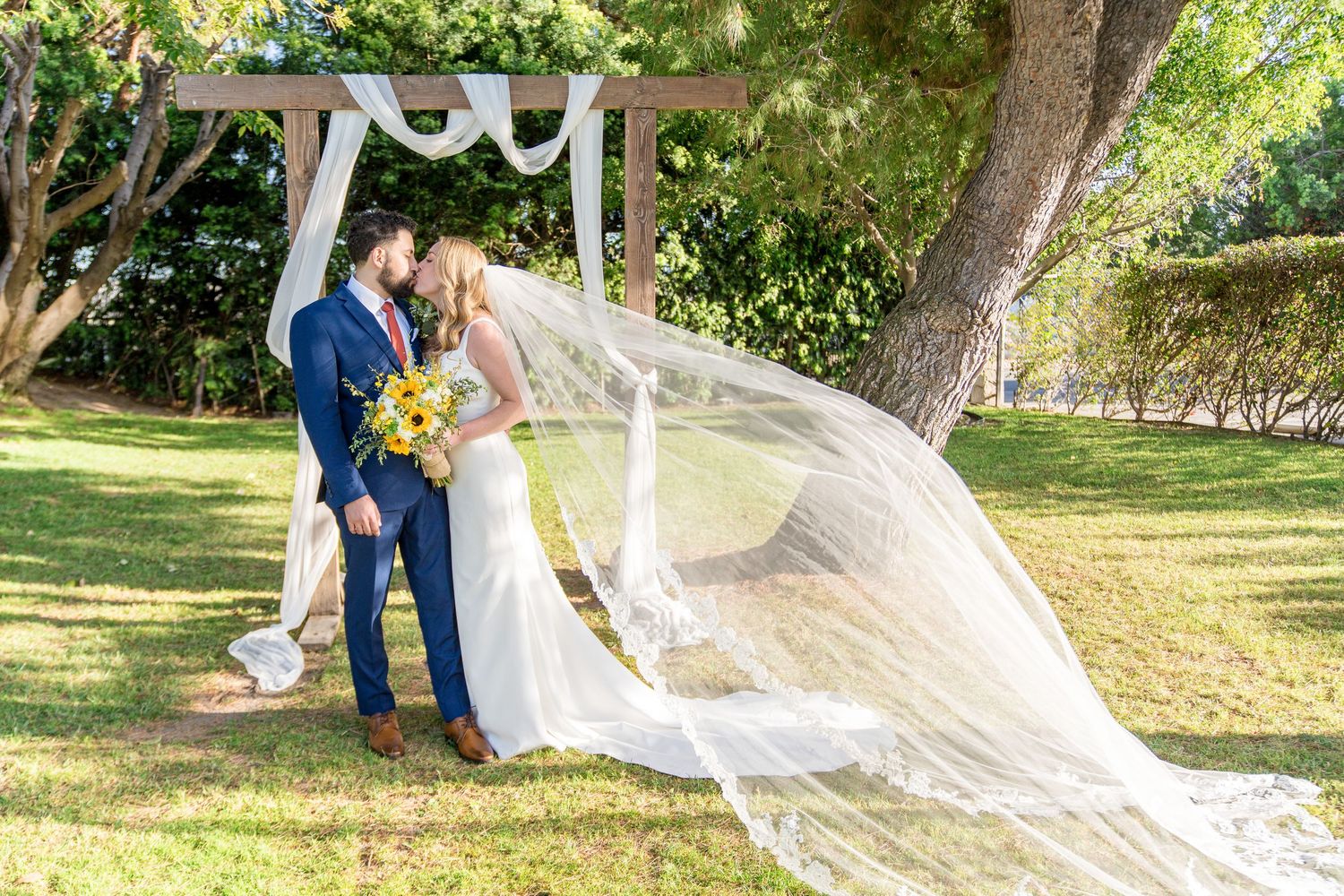 A long flowing veil catches the breeze as a couple stands together in a sunlit garden setting.
