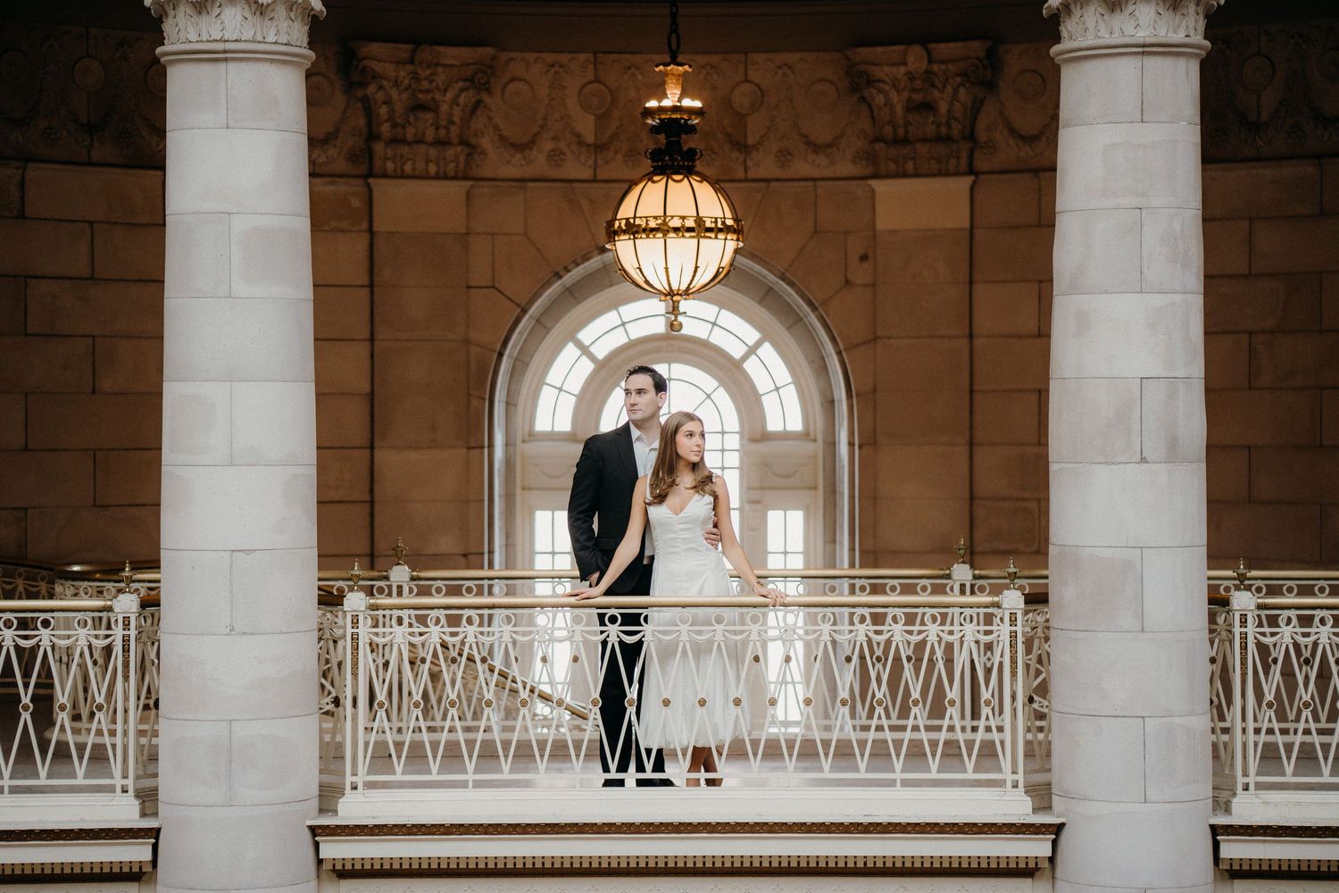 A couple stands together on an elegant balcony with ornate white railings and classical architecture featuring arches and columns.