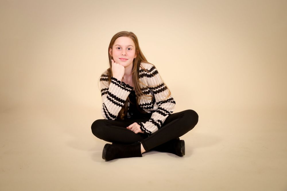 Casual studio portrait against beige backdrop showing relaxed cross-legged pose.
