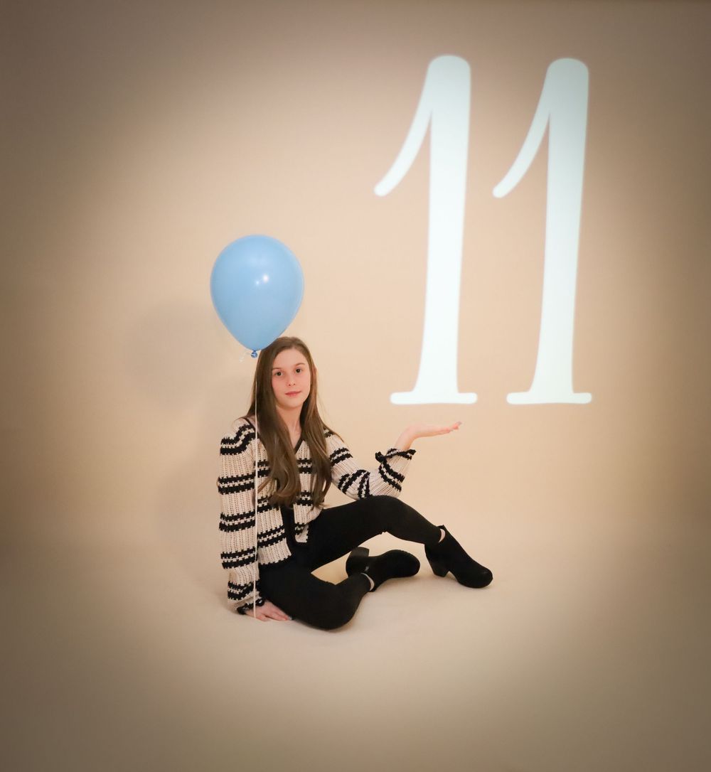 Person in striped sweater sitting with blue balloon next to large number 11 projection on wall.