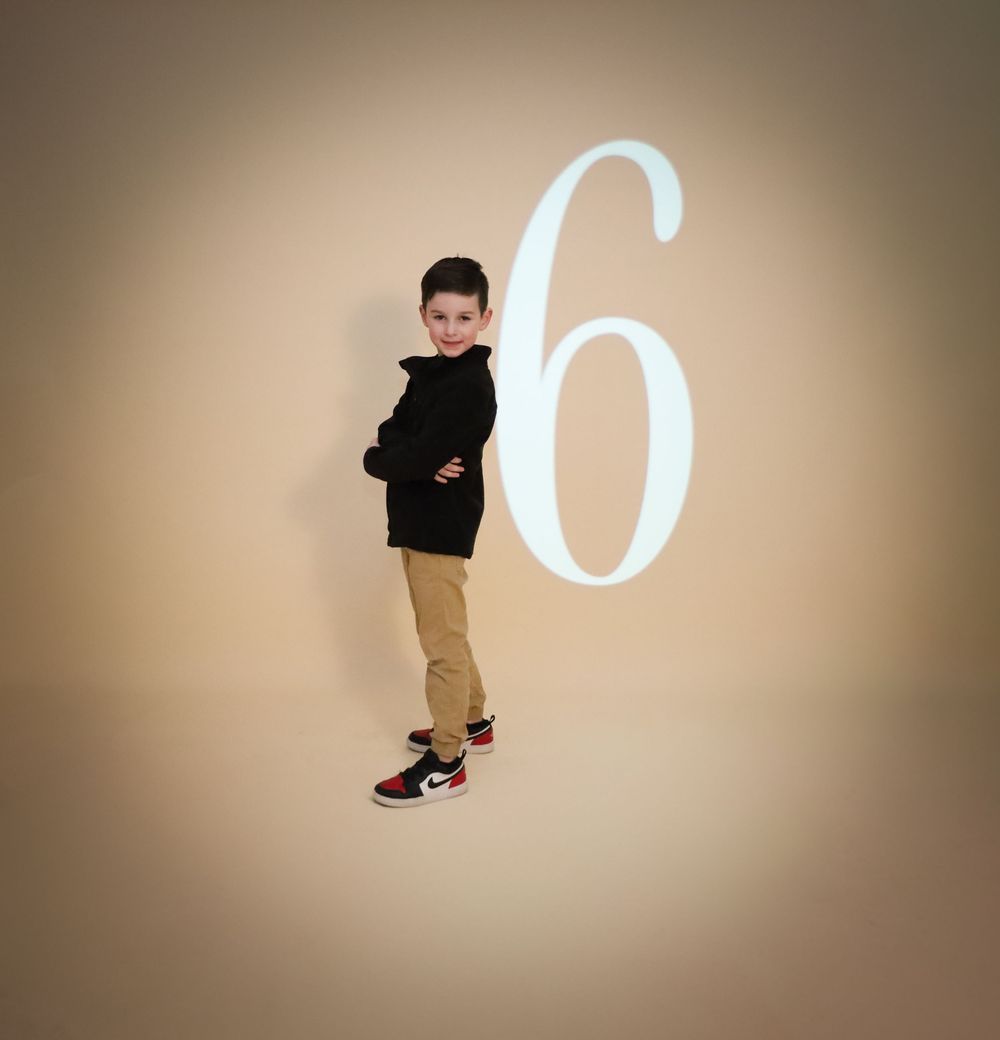Young person in black jacket poses confidently against wall with large number 6 projection.