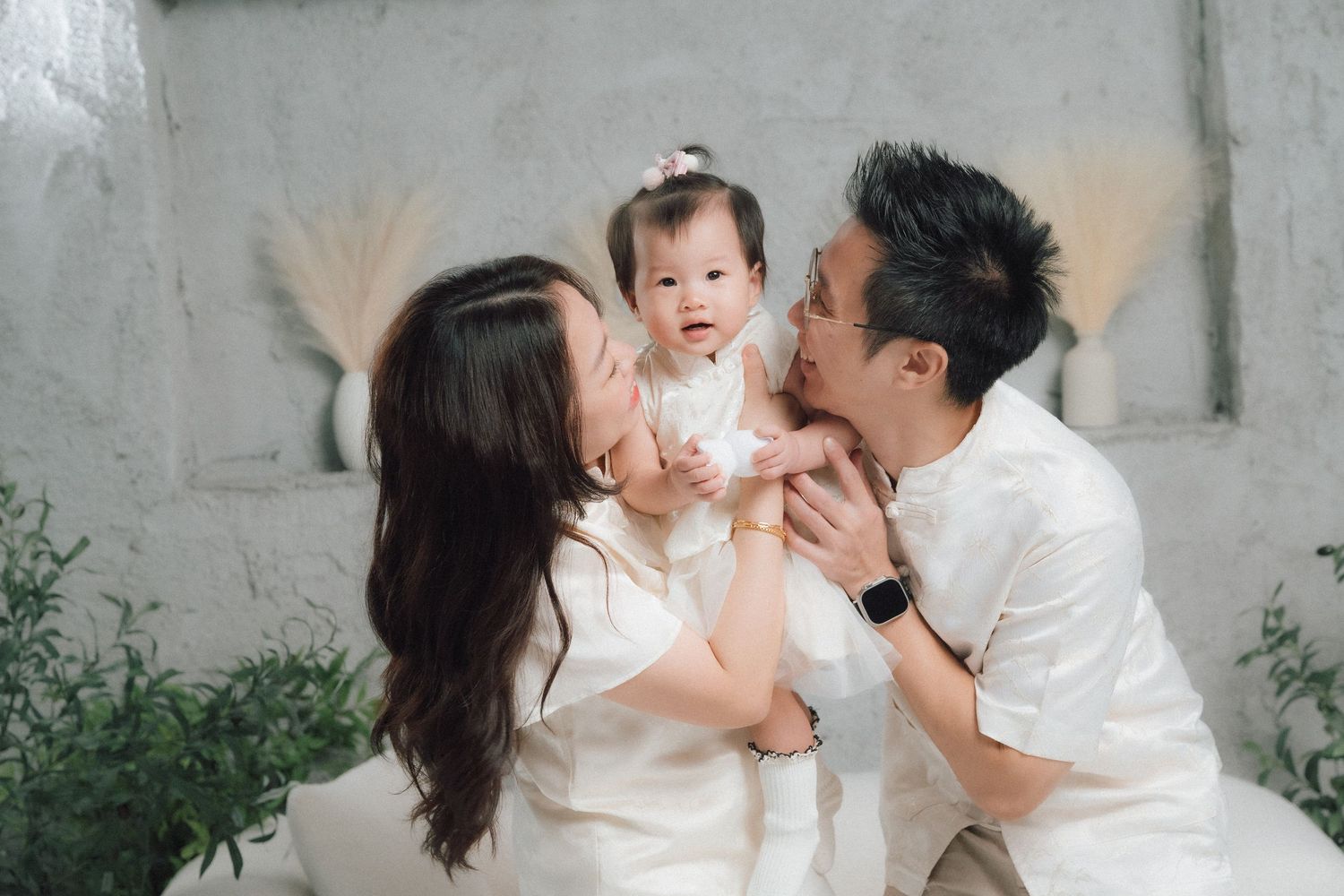 Family photo session in white outfits against grey wall with greenery.