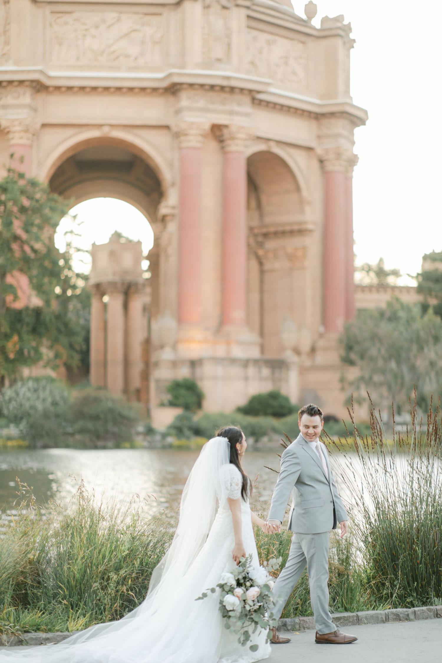 A couple in wedding attire stands by a pond near the Palace of Fine Arts in San Francisco at sunset.