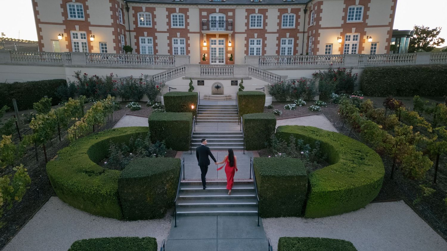 Two people walk down ornate garden steps leading to a grand historic mansion with manicured hedges at dusk.