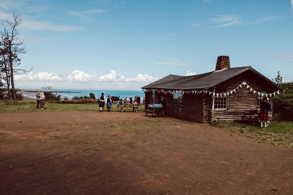 A rustic cabin with string lights sits on a grassy hilltop overlooking the ocean under a blue sky.