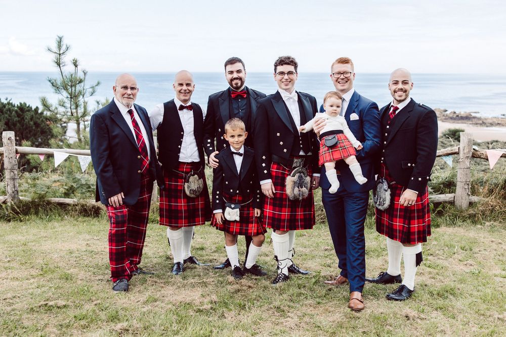 A group wearing traditional red and black kilts poses together outdoors against a scenic landscape backdrop.