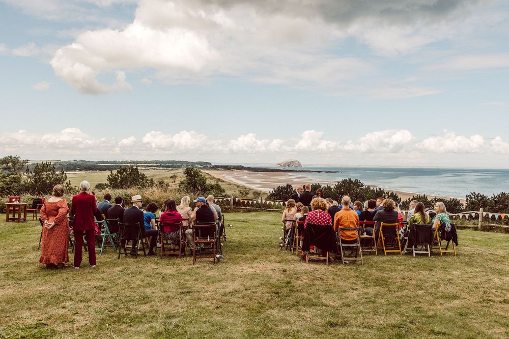 Panoramic view of outdoor wedding ceremony setup with wooden chairs facing the ocean under cloudy skies.