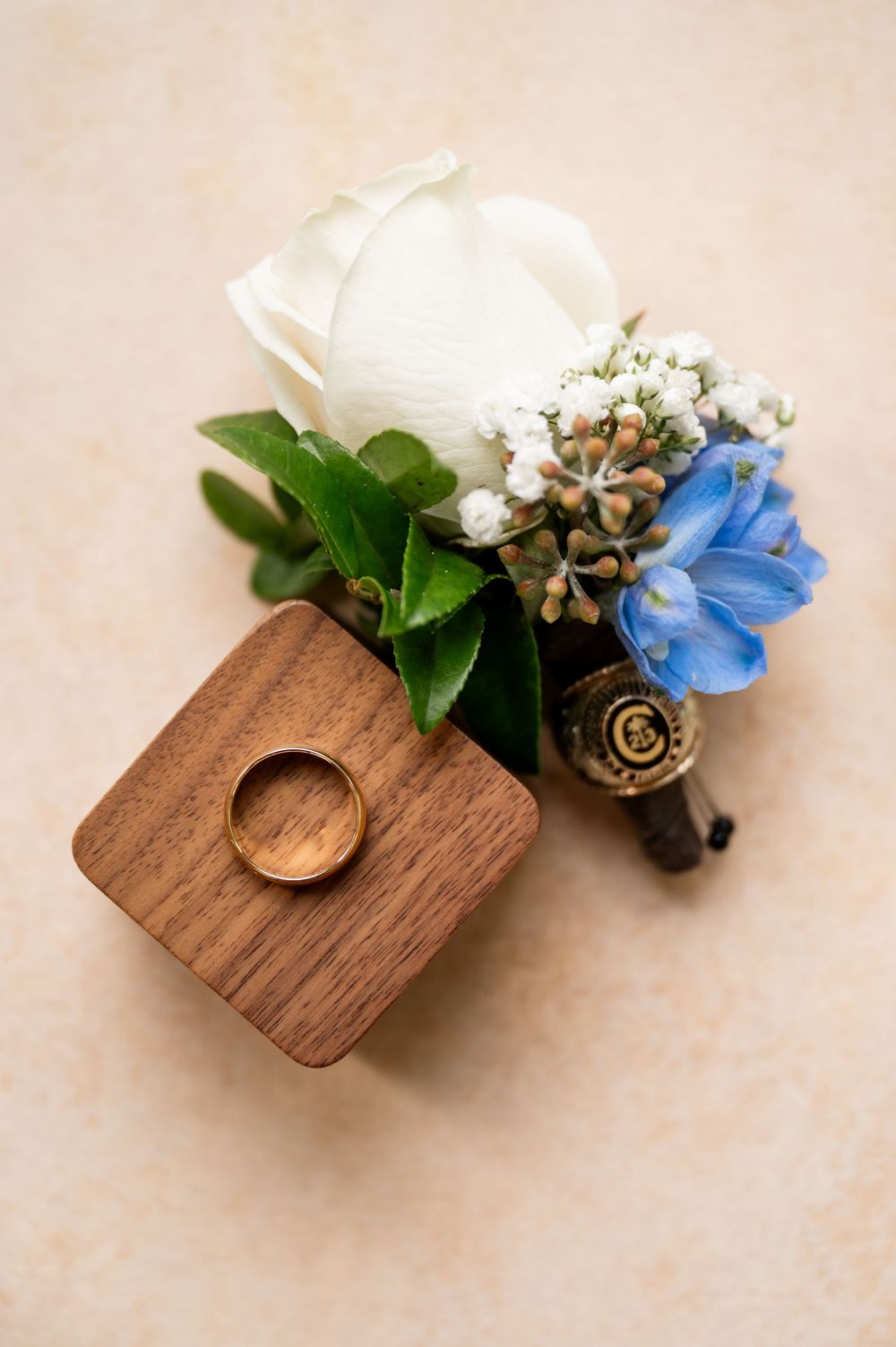 White rose boutonniere with blue accent flower on wooden ring holder.