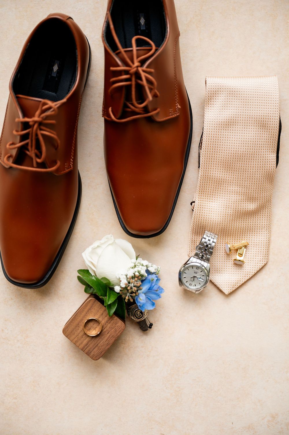 Brown leather dress shoes with tie, watch and boutonniere arranged on beige surface.