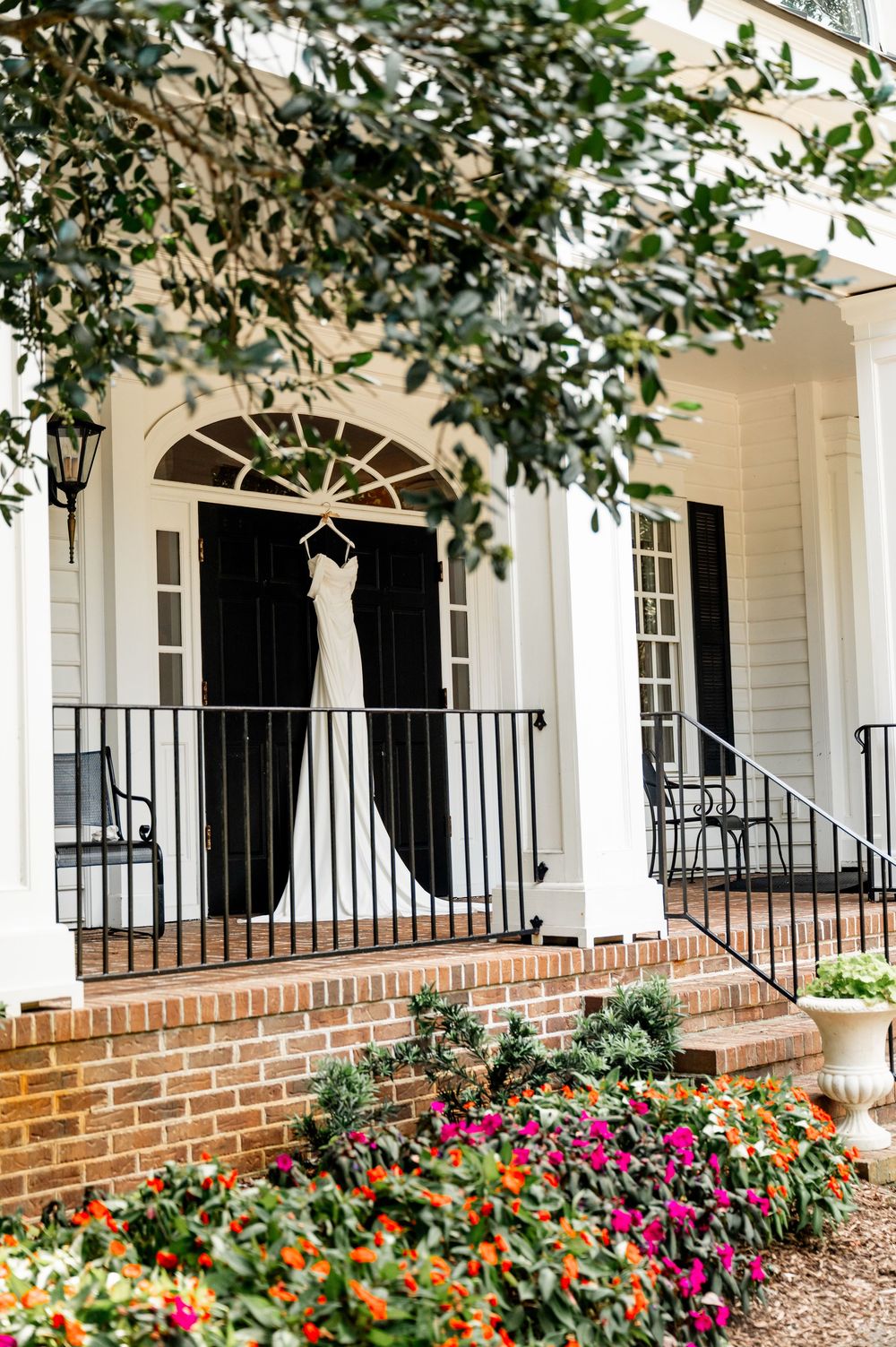 White wedding dress hanging on porch of classic colonial style house with colorful flower garden.