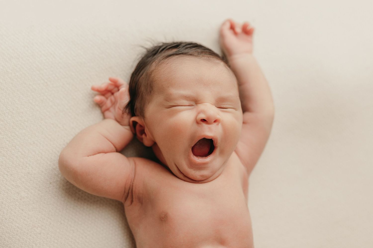 Newborn baby yawning and stretching while lying on beige backdrop during professional photo session.