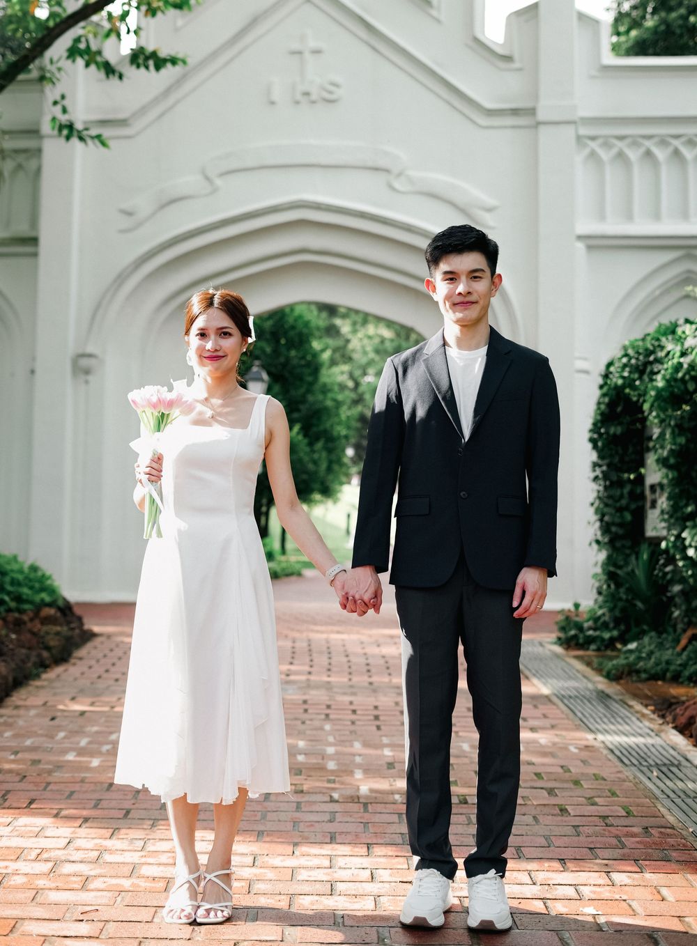 A newlywed couple holds hands on a brick path in front of a white church with Gothic architectural details.