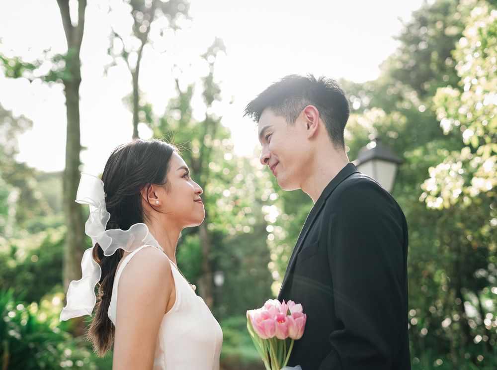 A couple in wedding attire share a tender moment in a sunlit garden setting with pink flowers.