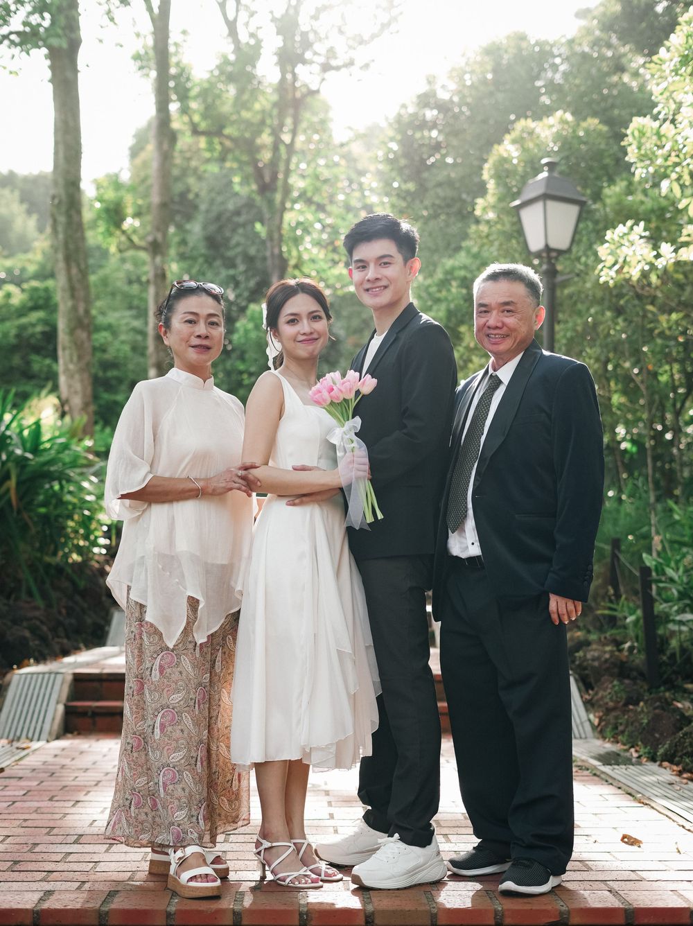 A wedding party poses outdoors on a brick pathway surrounded by lush greenery and trees on a sunny day.