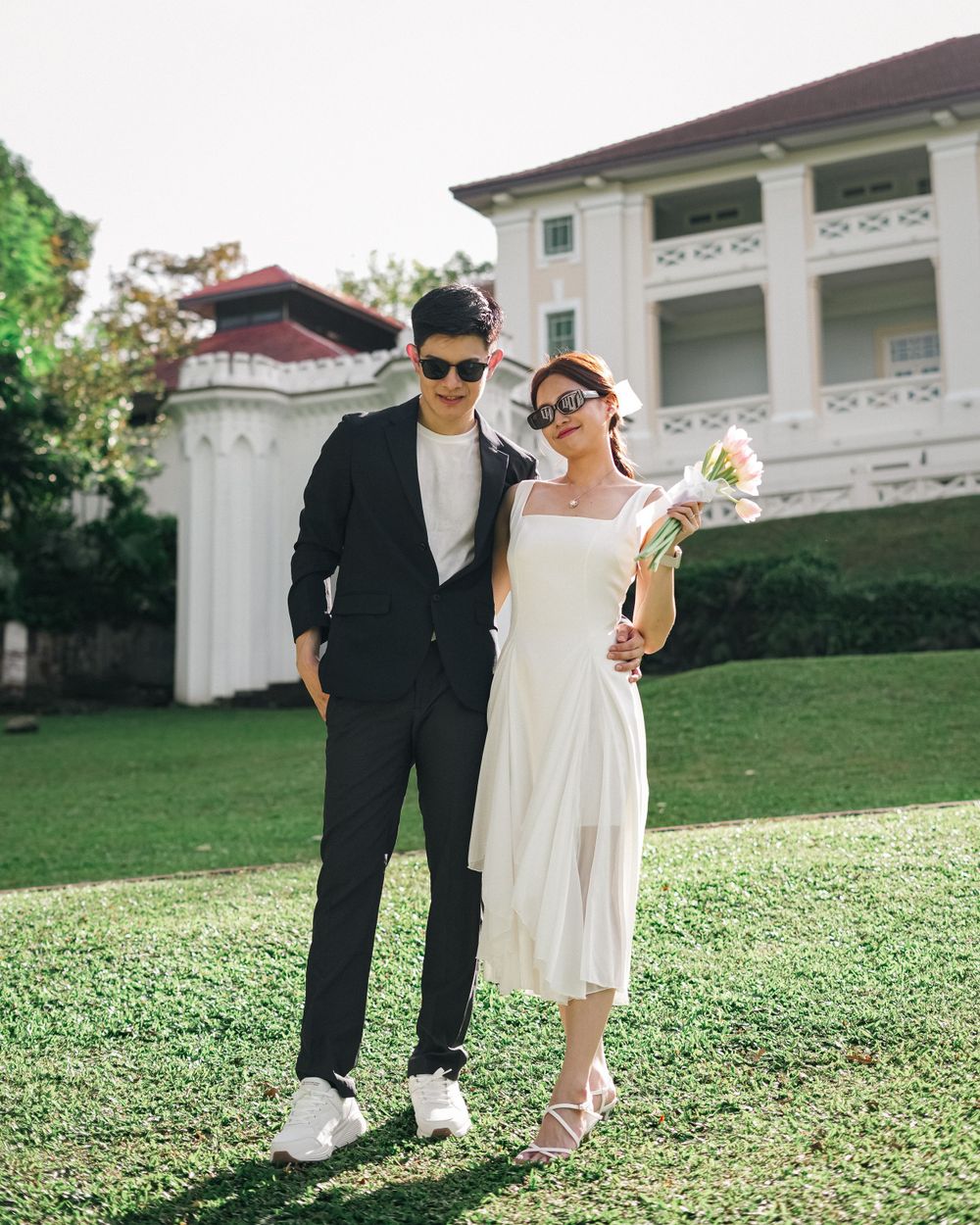 A couple in elegant contrasting outfits pose on a manicured lawn in front of a white colonial-style building.