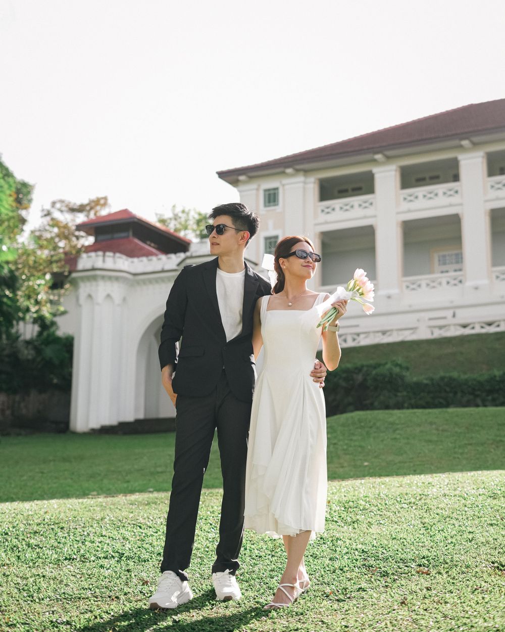 A stylish couple stands together on grass with a white mansion and garden wall in the background.