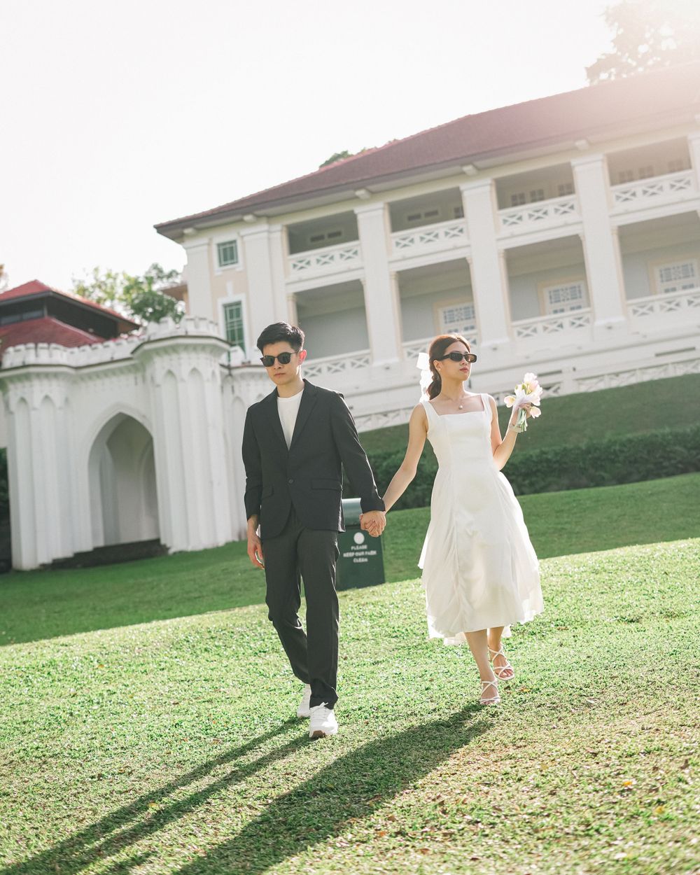 A couple in matching formal wear walks hand in hand on a grassy lawn with a white colonial building behind them.