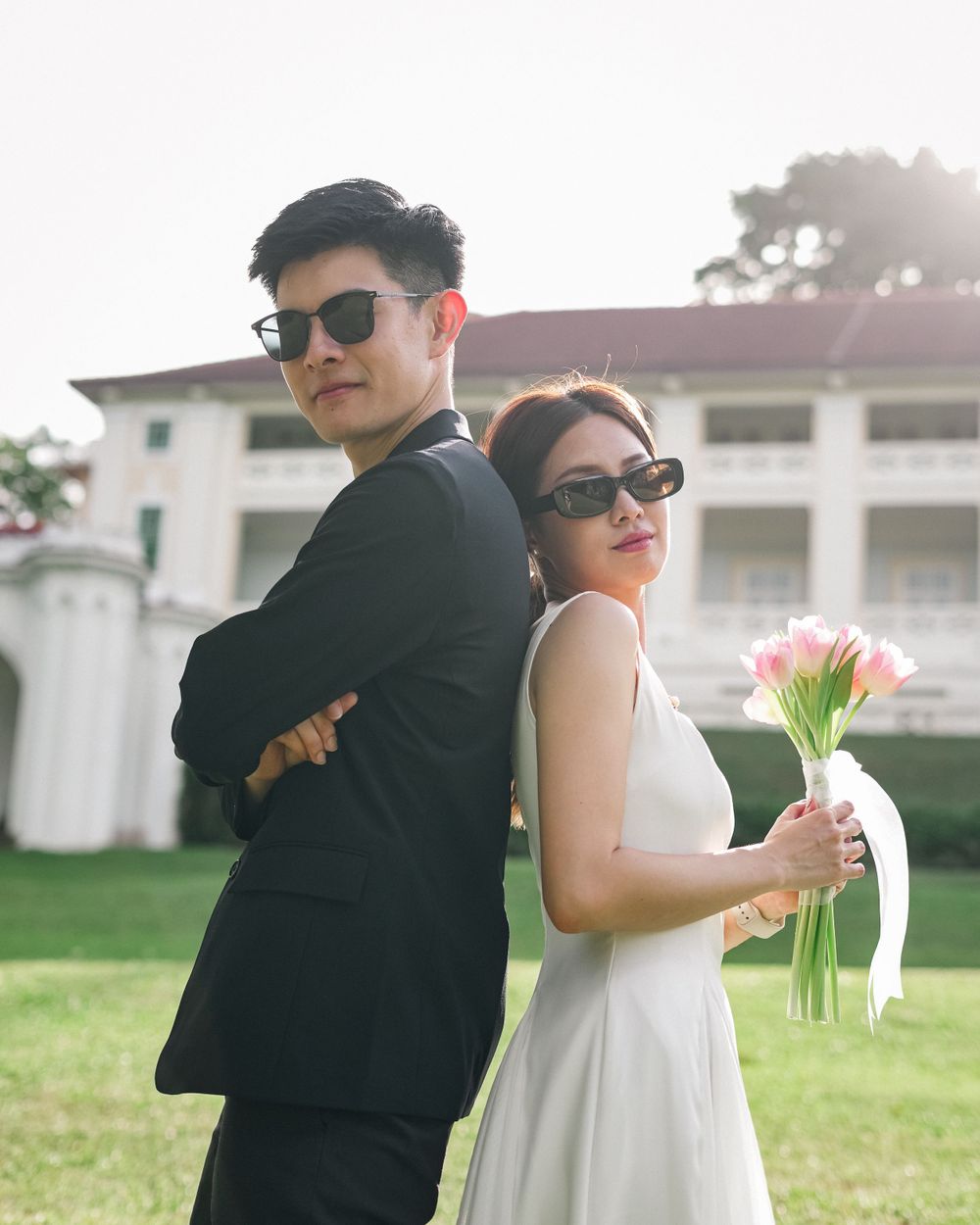 A couple in coordinated black and white outfits pose back-to-back with tulips on a sunny lawn.