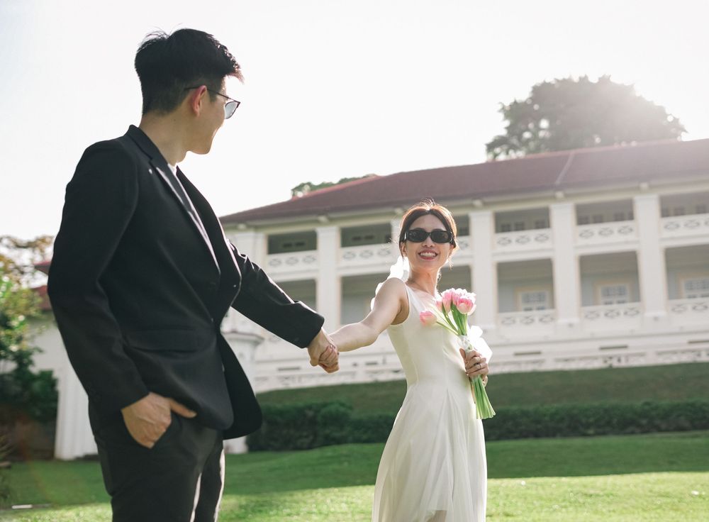 Two people in formal attire dancing playfully on a green lawn outside a white colonial-style mansion in the sunlight.