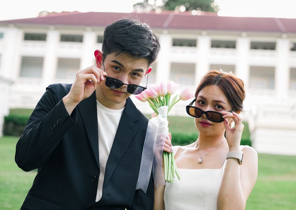 A playful couple adjusts their sunglasses while posing with flowers on a sunny lawn.