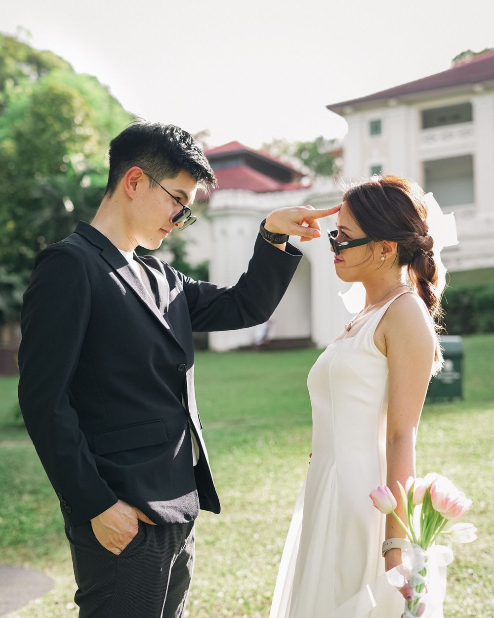 A couple in formal attire strikes a playful pose with hand gestures on a green lawn.