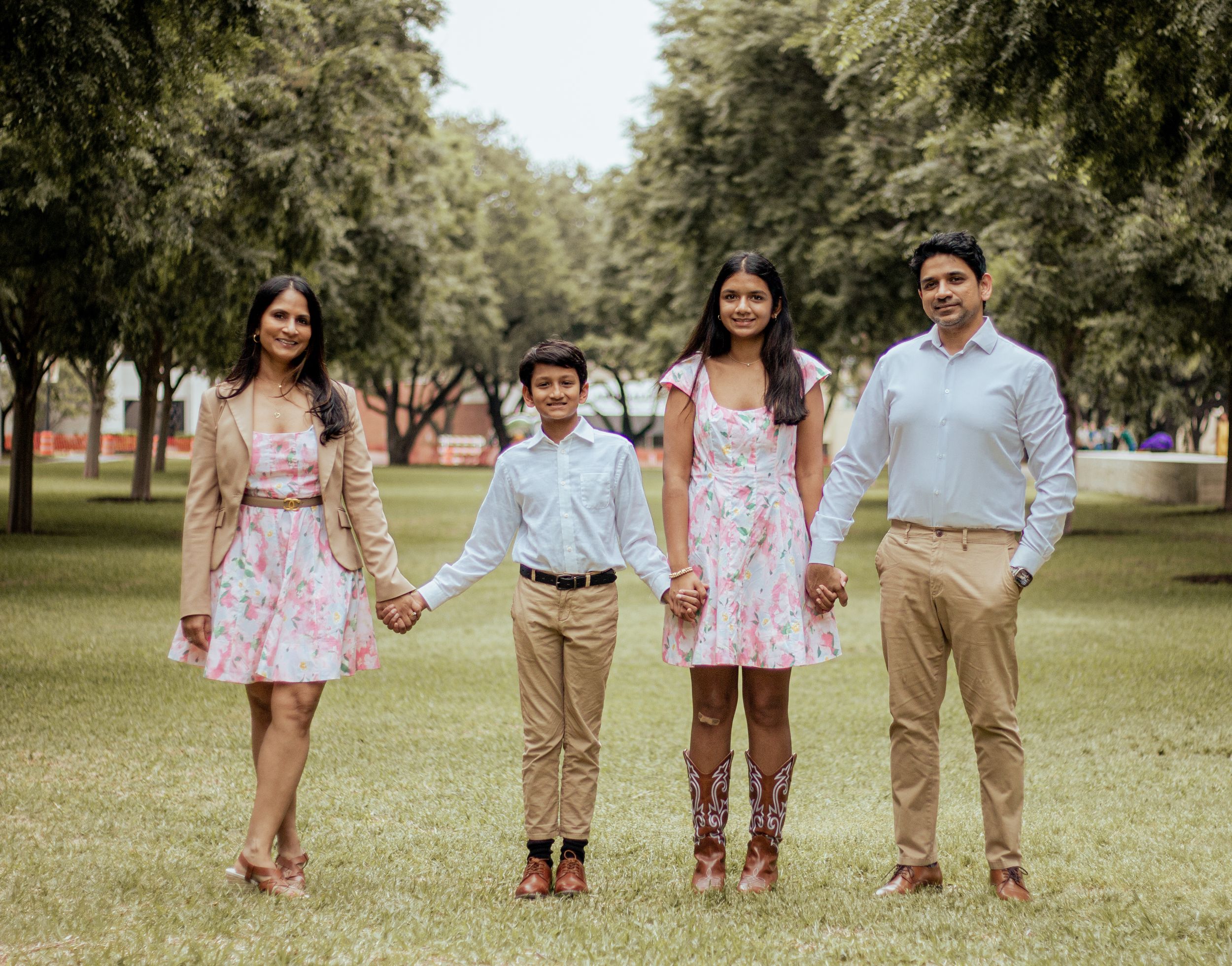 A family of four wearing coordinated pink and beige outfits stands together on green grass in a tree-lined park.