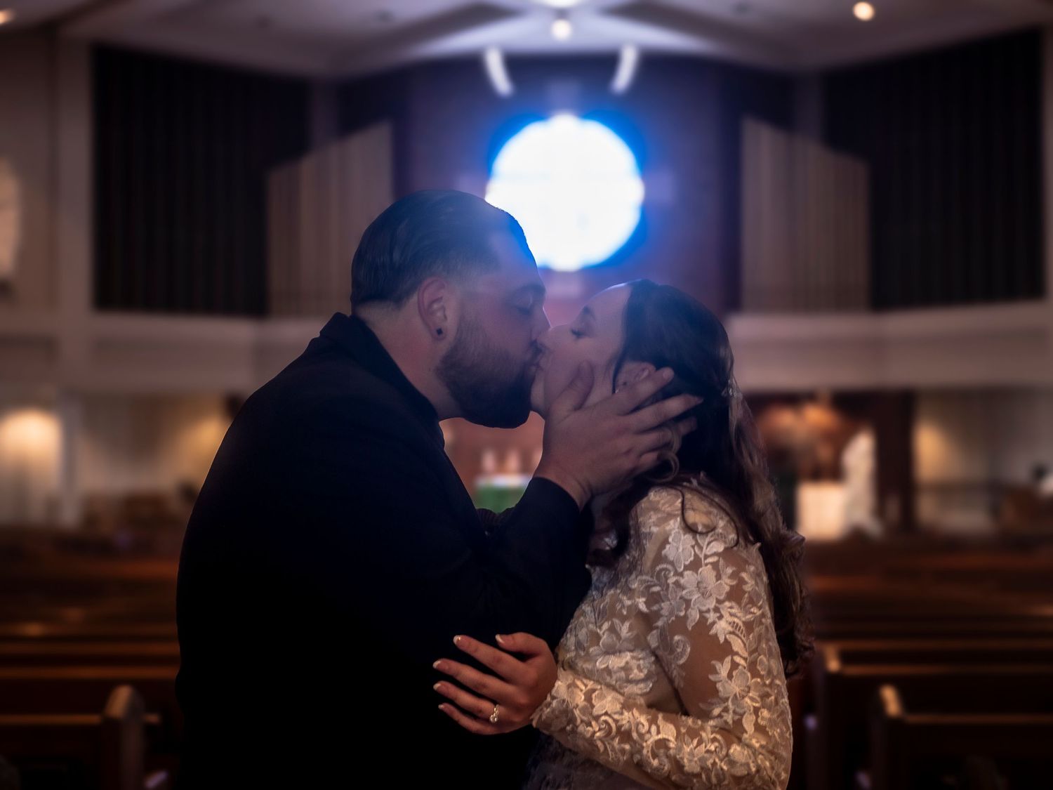 A couple shares a tender kiss with soft blue lighting in an empty church pew, the bride wearing an elegant lace gown.