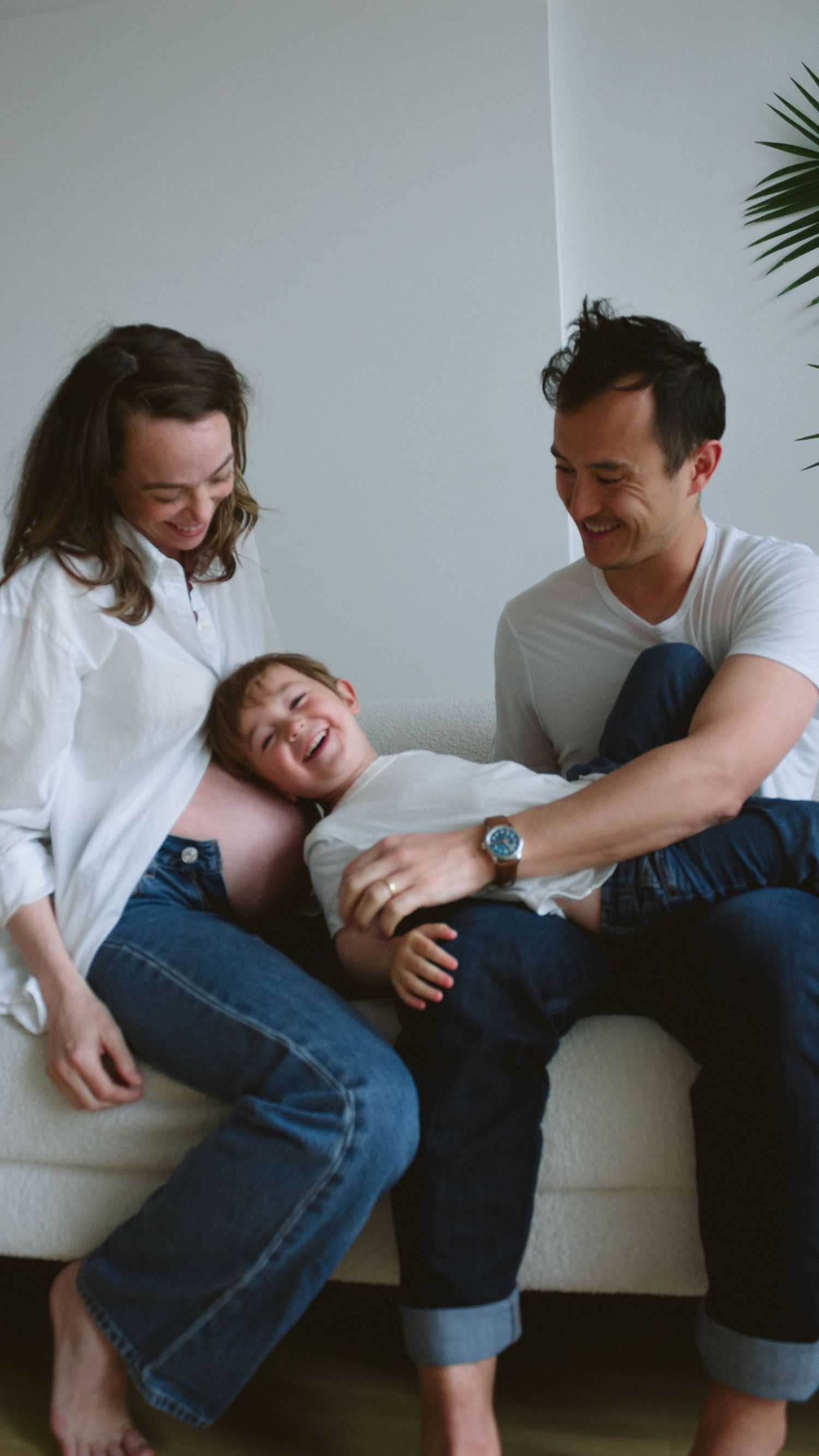 family sitting on couch with toddler between them, giggling on mom's pregnant belly