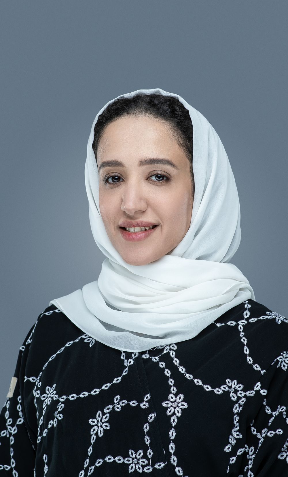 Young woman wearing a white hijab and black dress with embroidered floral chain pattern against gray background.