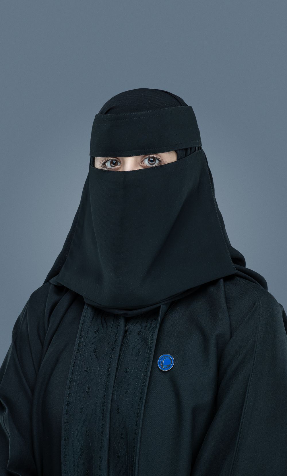 Studio portrait against gray background of someone wearing black niqab with bright blue button detail on garment.