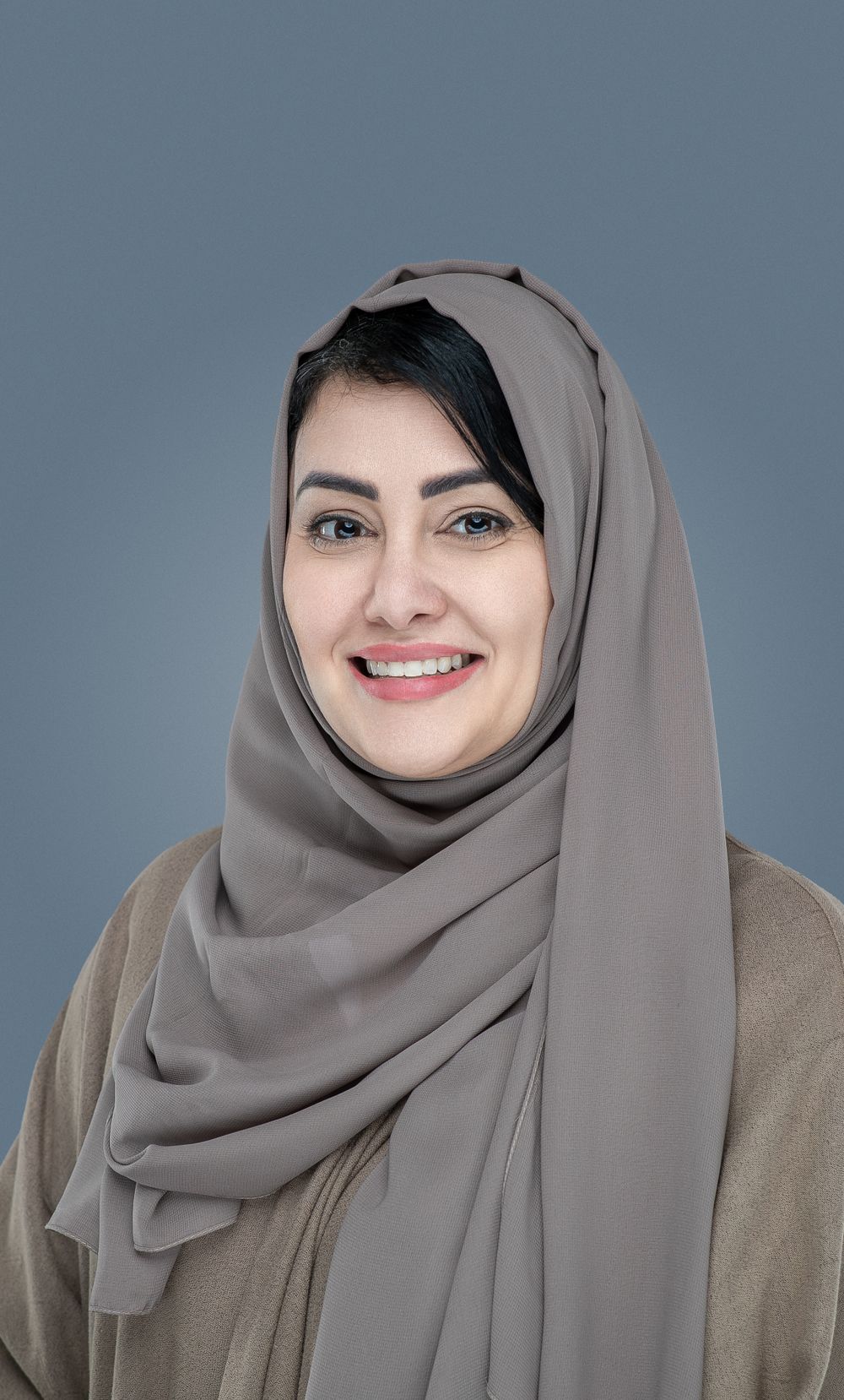 Professional studio portrait of someone wearing a gray hijab and beige top against a blue background.
