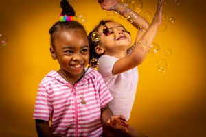 Two children in striped shirts jumping and playing with bubbles against yellow backdrop.