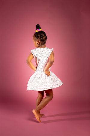 Back view of child in white dress posing playfully against pink backdrop.