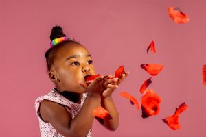 Child blowing red flower petals against pink background in white dress.