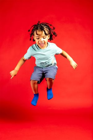 Energetic child wearing blue socks and shorts jumps joyfully against a vibrant red background.