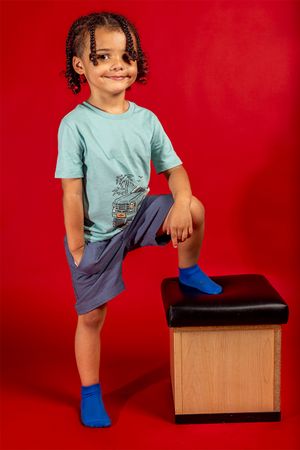 Child in light blue t-shirt and navy shorts poses with one foot on wooden box against red background.