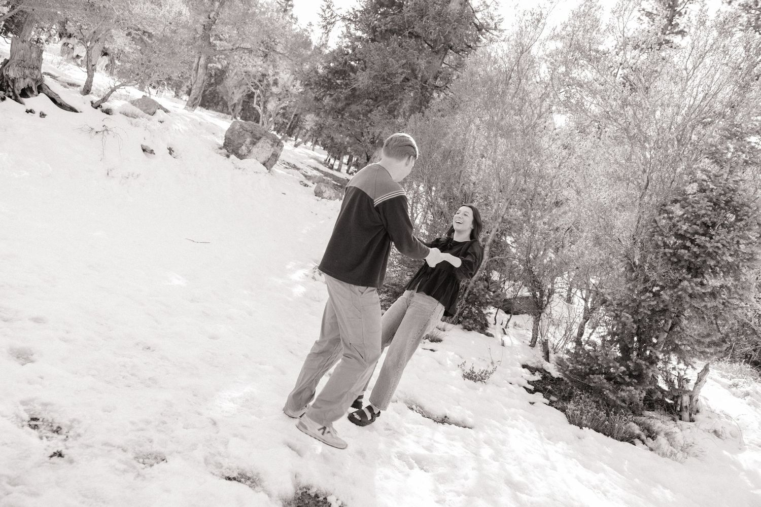 A couple stands together in a snowy winter scene with evergreen trees in the background.
