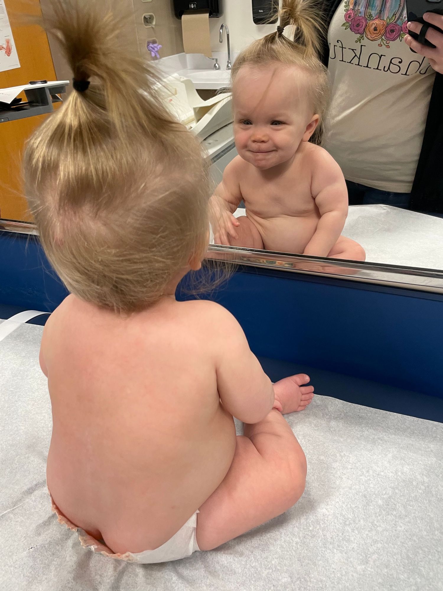 Baby in a diaper looks at their reflection in a mirror while sitting on an examination table.