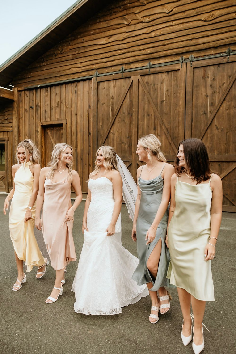 Bridal party wearing pastel color slip dresses walking together in front of rustic wooden barn doors.
