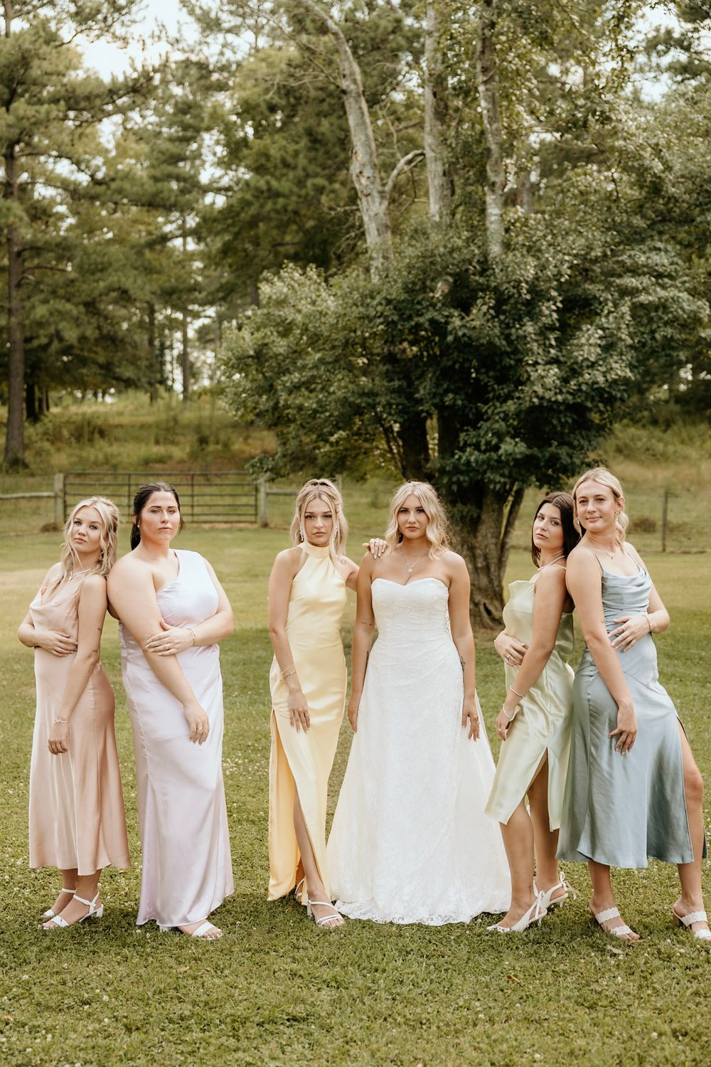 A bridal party in pastel dresses poses together outdoors on green grass with trees in the background.