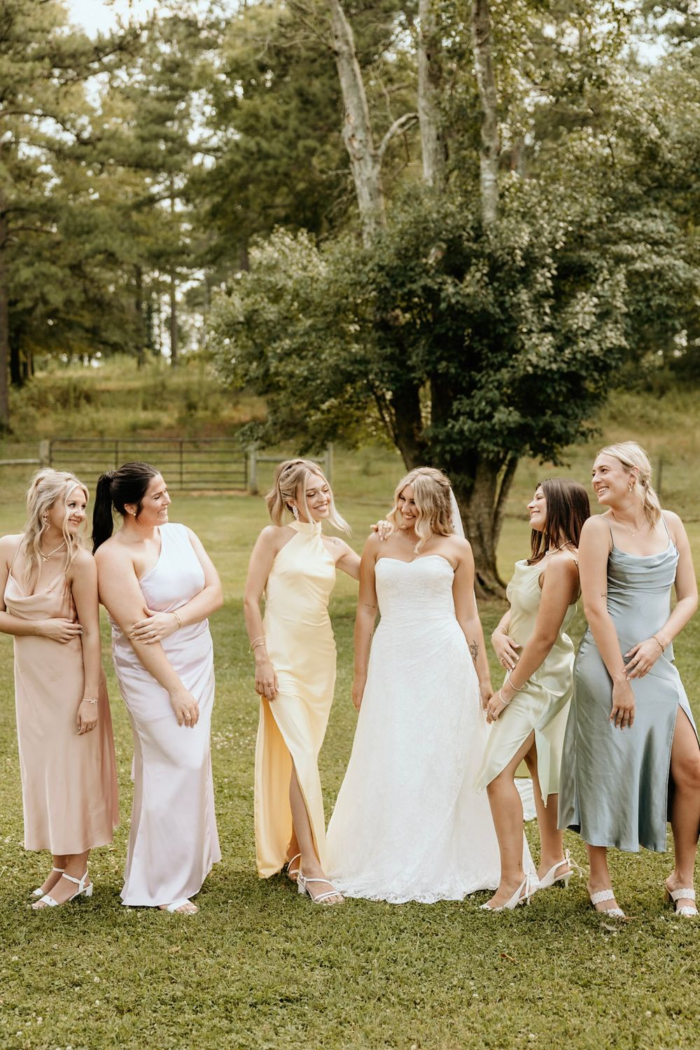 A wedding party stands on grass in a garden setting wearing pastel colored gowns in different styles and lengths.