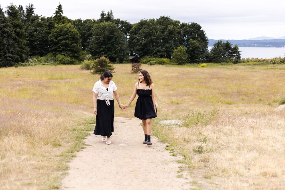 Two people walk along a dirt path through a grassy field with evergreen trees and water visible in the distance.