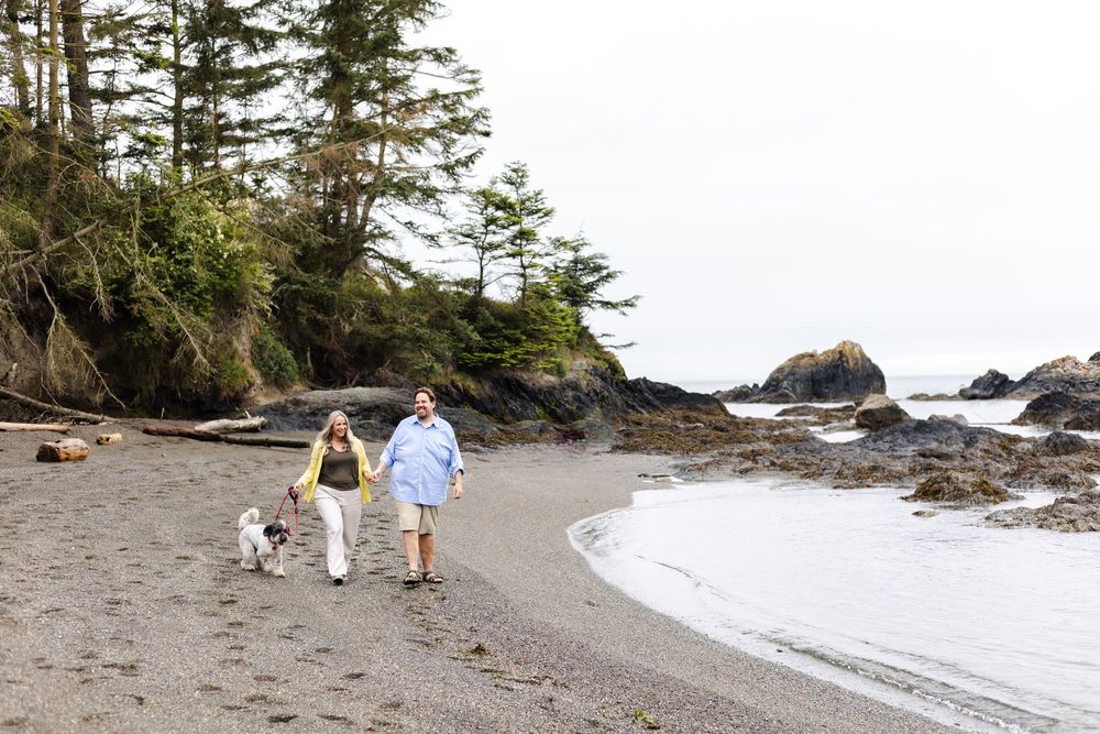 Two people and a dog walking along a rocky Pacific Northwest beach with evergreen trees and coastal rocks in the background.