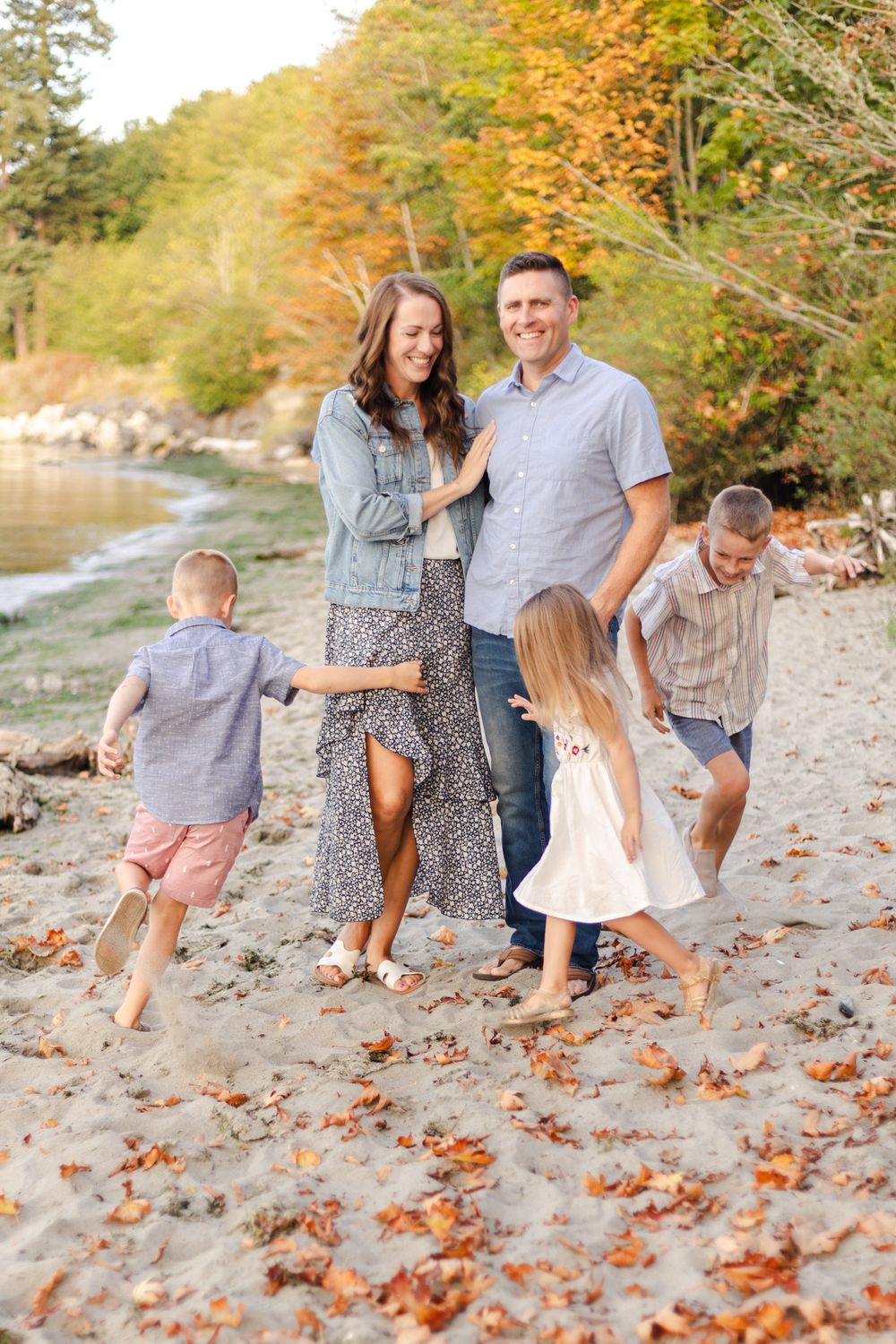 A family plays together on a beach surrounded by autumn leaves and trees during a fall photoshoot session.