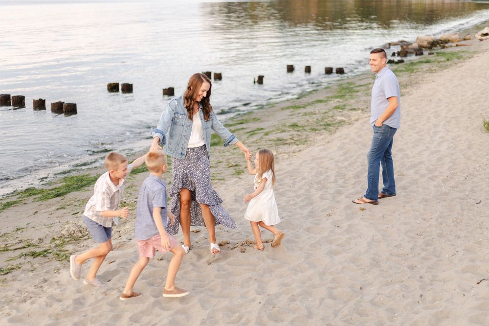 A group walks along a sandy beach shoreline with water and wooden posts visible in the background.