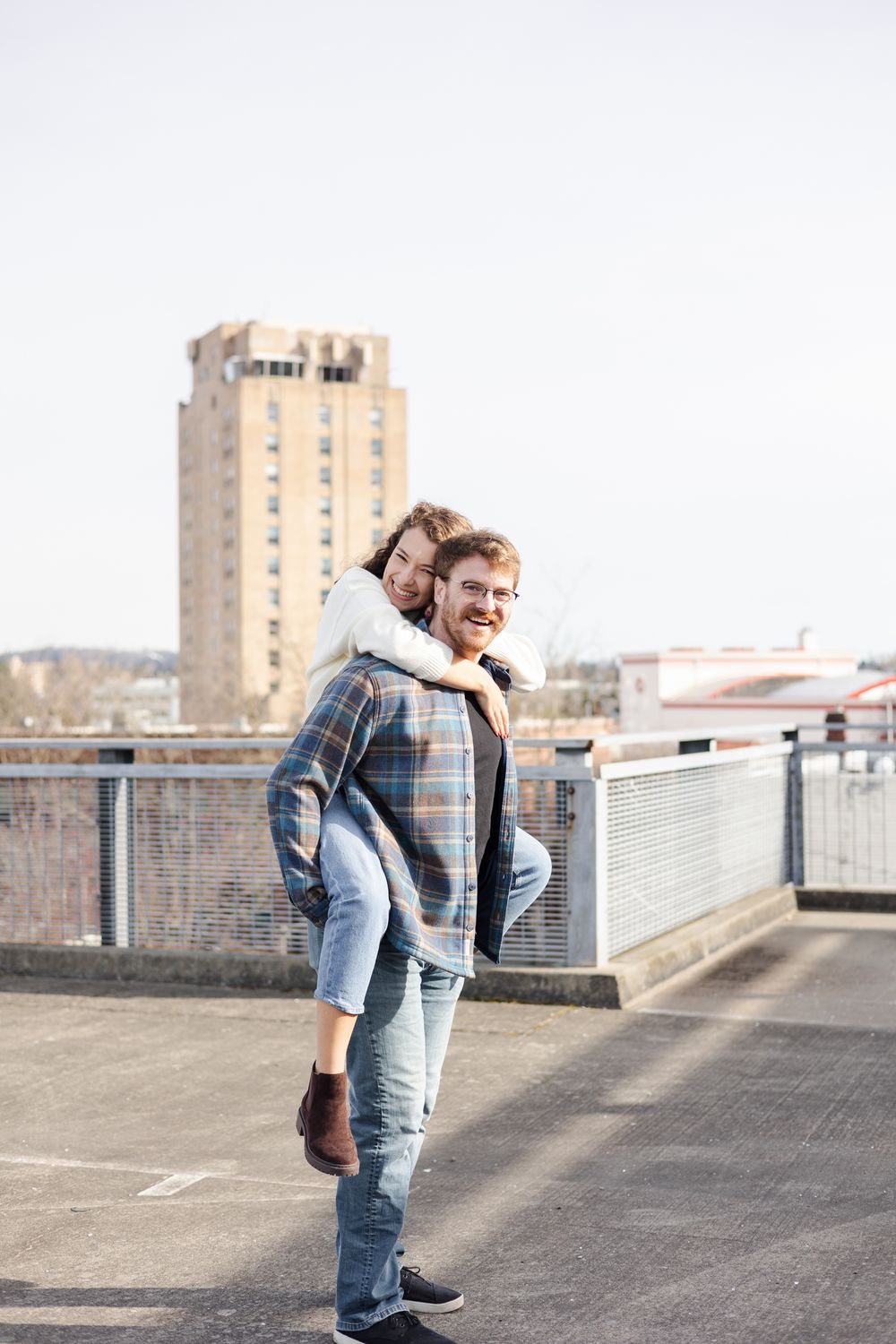 Person in plaid jacket and jeans poses casually on urban rooftop with tall building in background.