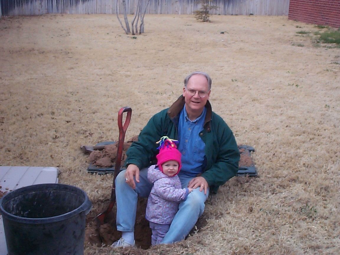 A person in a green jacket sits with a small child in pink winter clothing on brown grass near a black bucket.
